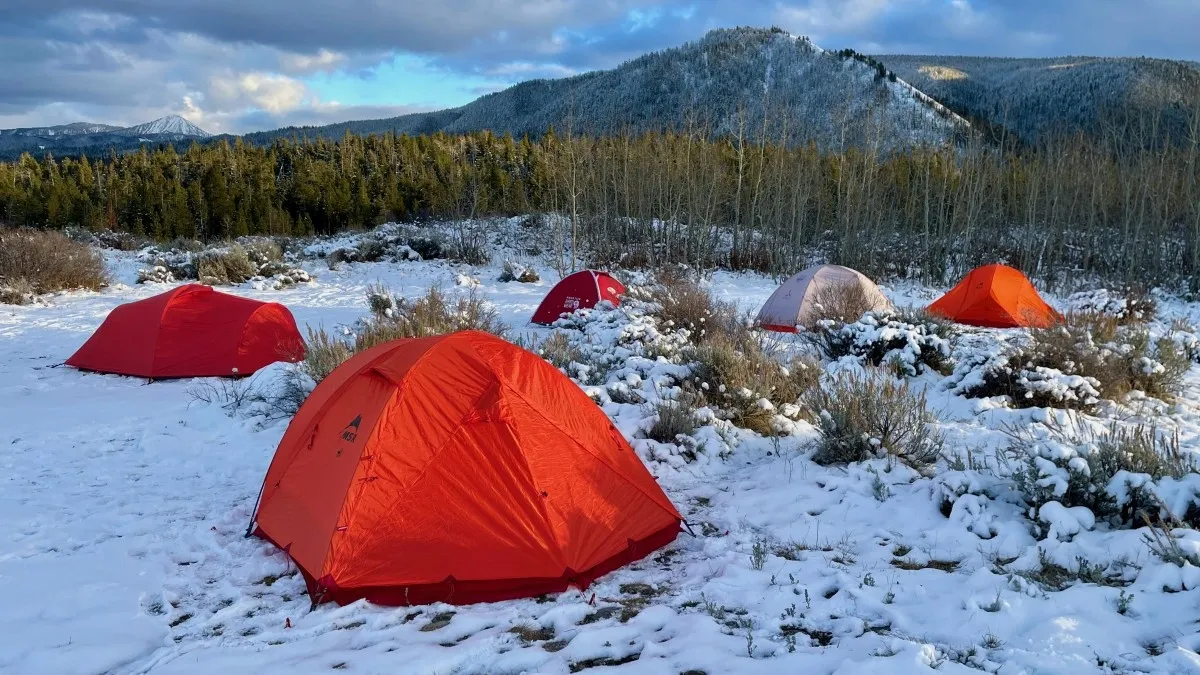 Six colorful 4-season tents pitched on a snowy field with forest and mountain backdrop under a cloudy sky.