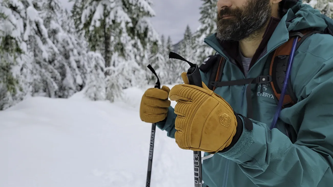 Person wearing tan insulated winter gloves while holding trekking poles in a snowy forest setting.